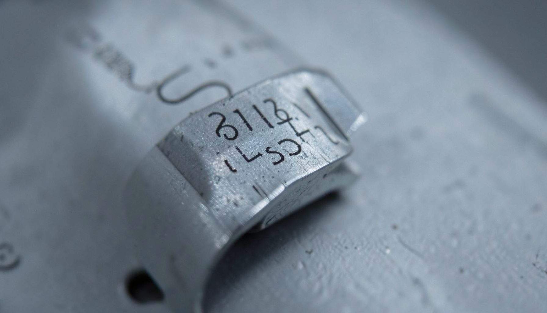 Small metal component with a fine dark marked code on a curved surface, cool gray tones suggesting industrial precision work